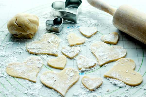 sugar cookie heart-shaped dough cutouts on top of a rolling mat with a rolling pin and cookie cutters on the side