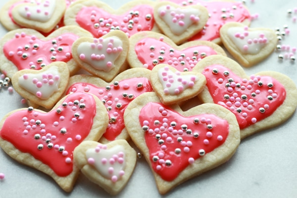 Heart-shaped sugar cookies in various sizes with pink and white glaze and decorative sprinkles stacked on top of a marble surface.