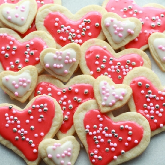 Heart-shaped sugar cookies in various sizes with pink and white glaze and decorative sprinkles stacked on top of a marble surface.