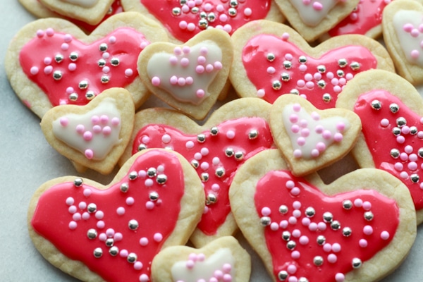 Heart-shaped sugar cookies in various sizes with pink and white glaze and decorative sprinkles stacked on top of a marble surface.