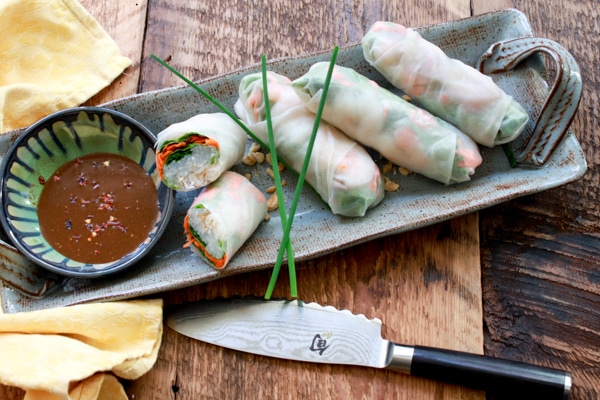 Fresh Vietnamese shrimp spring rolls on a long gray platter with a peanut sauce on the side on top of a wooden board with a knife and napkin.