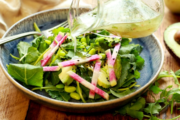 avocado-edamame kale salad in a blue bowl with dressing being poured from a glass dispenser