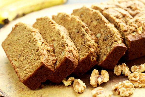 Sliced banana bread on a wooden board with walnuts and bananas on the side