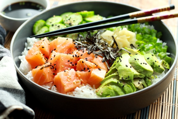 Salmon sushi bowl featuring chunks of sushi-grade salmon, sushi rice, sliced avocados, cucumbers, and seaweed strips on a bamboo mat with chopsticks.