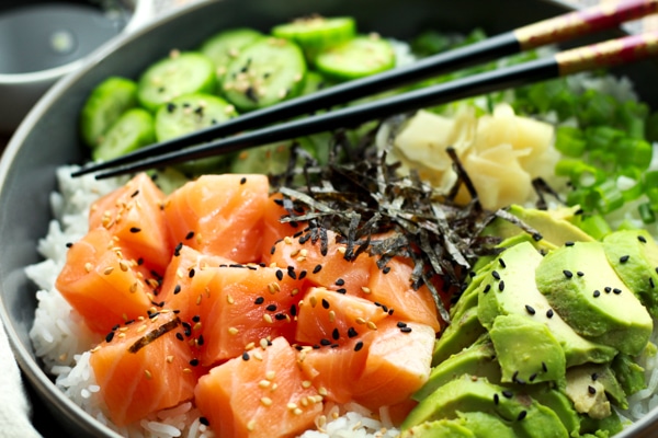 Salmon sushi bowl featuring chunks of sushi-grade salmon, sushi rice, sliced avocados, cucumbers, and seaweed strips with chopsticks.