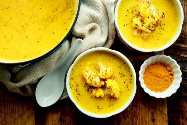 vibrant cauliflower soup in two white bowls on a wooden board with turmeric powder along side