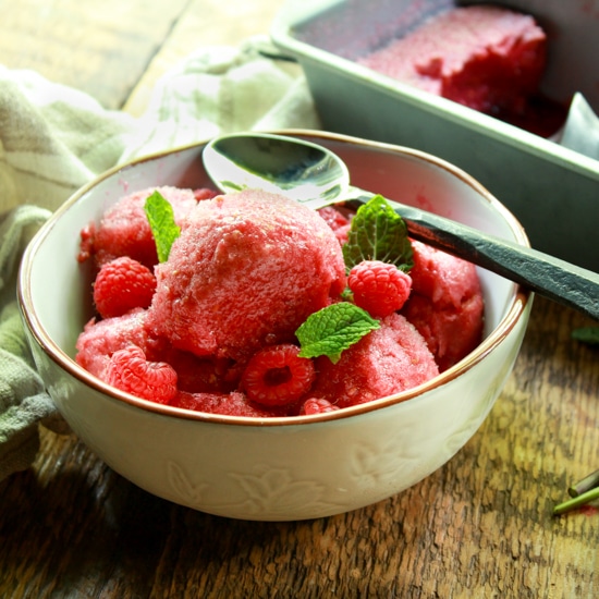 a white bowl of raspberry sorbet and spoon in a white bowl on a wooden board with a metal pan filled with sorbet