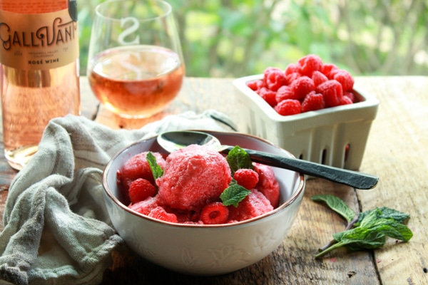 a white bowl of raspberry sorbet with a spoon on a wooden board with a bottle and glass of Gallivant Rosé and fresh raspberries on the side
