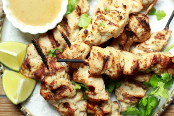 Tender satay chicken pieces cooked on wooden skewers on a white plate with a side of peanut sauce and fresh lime wedges