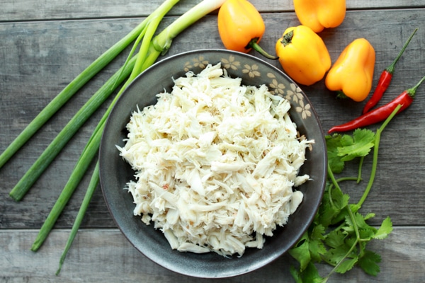 A bowl of lump crab meat on a wooden board with cilantro, peppers, and scallions on the side