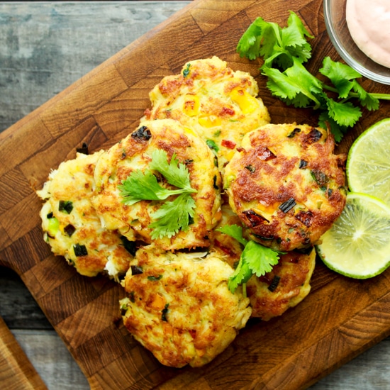 Mini crab cakes stacked on a wooden board with a side of aioli dipping sauce and fresh limes