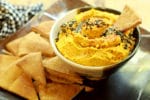 a small bowl of roasted carrot and ginger hummus with a pita chip inserted and pita chips on the side