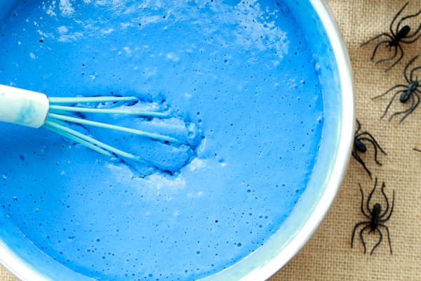 blue pancake batter in a large bowl with a whisk inside and black spiders crawling on a burlap surface