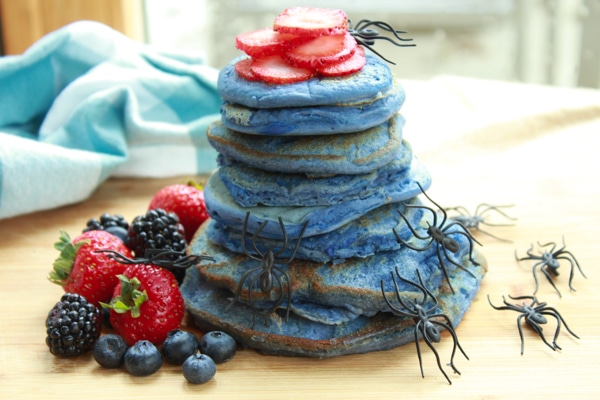 a stack of blue pancakes on a wooden board with vibrant berries on the side and plastic spiders crawling up the stack