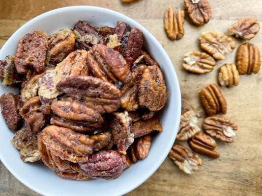 A white bowl filled with golden, spiced pecans and loose raw pecans on the side on a wooden board.