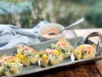 A long gray serving platter with baked wonton cups filled with crab salad and a dollop of colorful aioli on top on a wooden board and a small white bowl of aioli behind it.