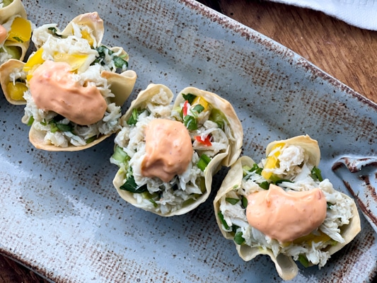 A long gray serving platter with baked wonton cups filled with crab salad and a dollop of aioli on top sitting on top of a wooden board.