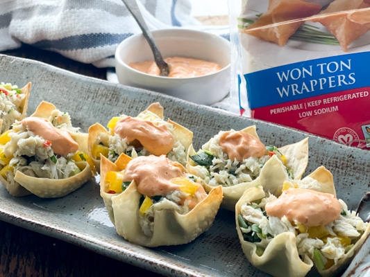 A gray serving platter with wonton cups filled with crab salad and a dollop of aioli on top with the package of wonton wrappers behind it.