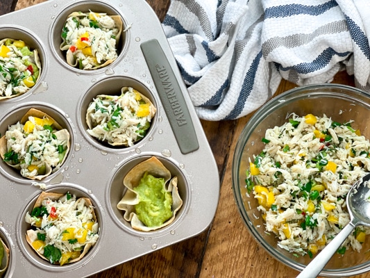 An aluminum muffin tray filled with baked wontons filled with crab salad and guacamole and a clear glass bowl with crab salad on the side on top of a wooden board. on a wooden board.