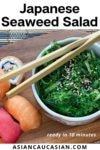 A bowl filled with Japanese seaweed salad with black chopsticks on top and assorted sushi on the side, on a wooden board.