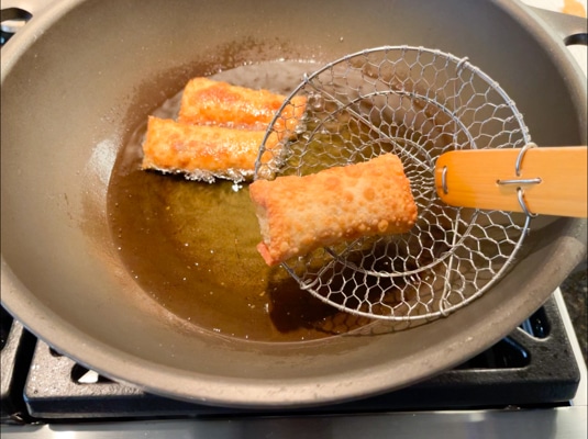 Golden fried spring rolls being lifted out of a large wok with a slotted spoon.