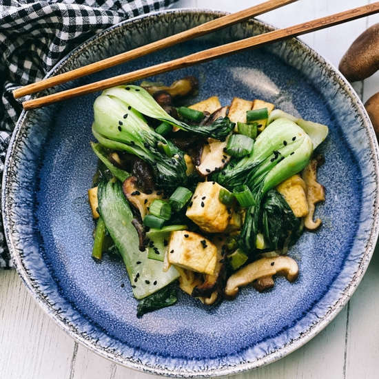 Stir-fry miso shiitake mushrooms, tofu, and baby bok choy on a blue plate with wooden chopsticks, and fresh shiitake mushrooms on the side.