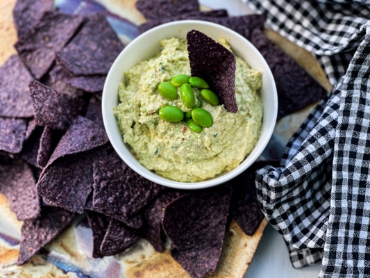 Goat cheese edamame dip in a small white bowl with edamame pods and blue corn tortilla chips on the side.
