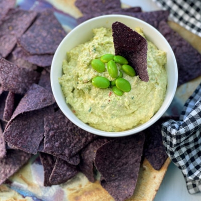 Goat cheese edamame dip in a small white bowl with edamame pods and blue corn tortilla chips on the side.