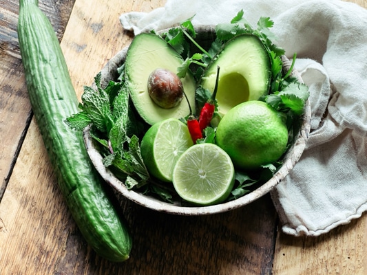 Ingredients in a wooden bowl for cucumber avocado soup: limes, avocado, chili peppers, green herbs and a whole cucumber.