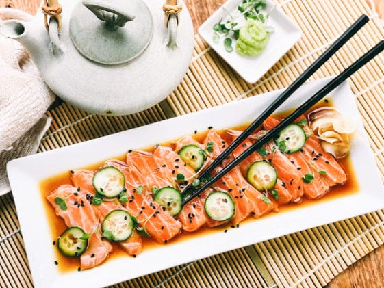 Slices of fresh sashimi salmon on a white platter with sliced cucumbers on top and sprinkles of black sesame seeds, with black chopsticks, a Japanese tea kettle, and a small white bowl of green wasabi on the side.