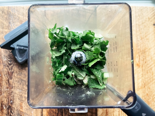 Fresh herbs in a blender on top of a wooden board.