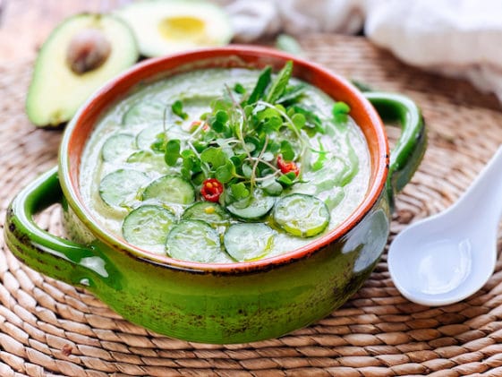 A bowl filled with spicy cucumber avocado soup topped with sliced cucumbers and herbs with a sliced avocado and a white spoon on the side.