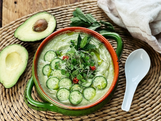 A bowl filled with spicy cucumber avocado soup topped with sliced cucumbers and herbs with a sliced avocado and a white spoon on the side.