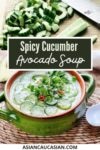 A bowl filled with spicy cucumber avocado soup topped with sliced cucumbers and herbs with a sliced avocado and a white spoon on the side.