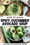 A bowl filled with spicy cucumber avocado soup topped with sliced cucumbers and herbs with a sliced avocado and a white spoon on the side.