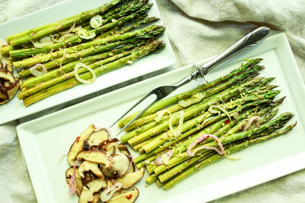 Roasted asparagus and shiitake mushrooms on two white trays with a serving fork on the side.