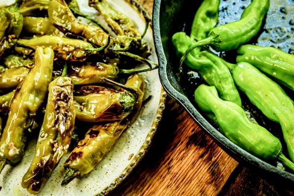 A plateful of blistered shishito peppers and raw peppers in a cast iron pan