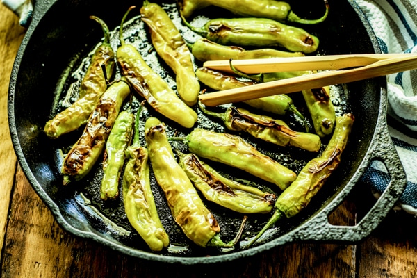 Blistered shishito peppers in a cast iron pan with wooden tongs inserted.