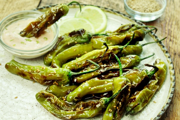 Blistered shishito peppers on a white plate with lime wedges, aioli dipping sauce, and sesame seeds on the side.