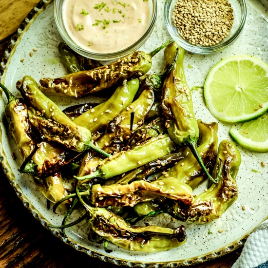 Blistered shishito peppers on a white plate with lime wedges, aioli dipping sauce, and sesame seeds on the side.