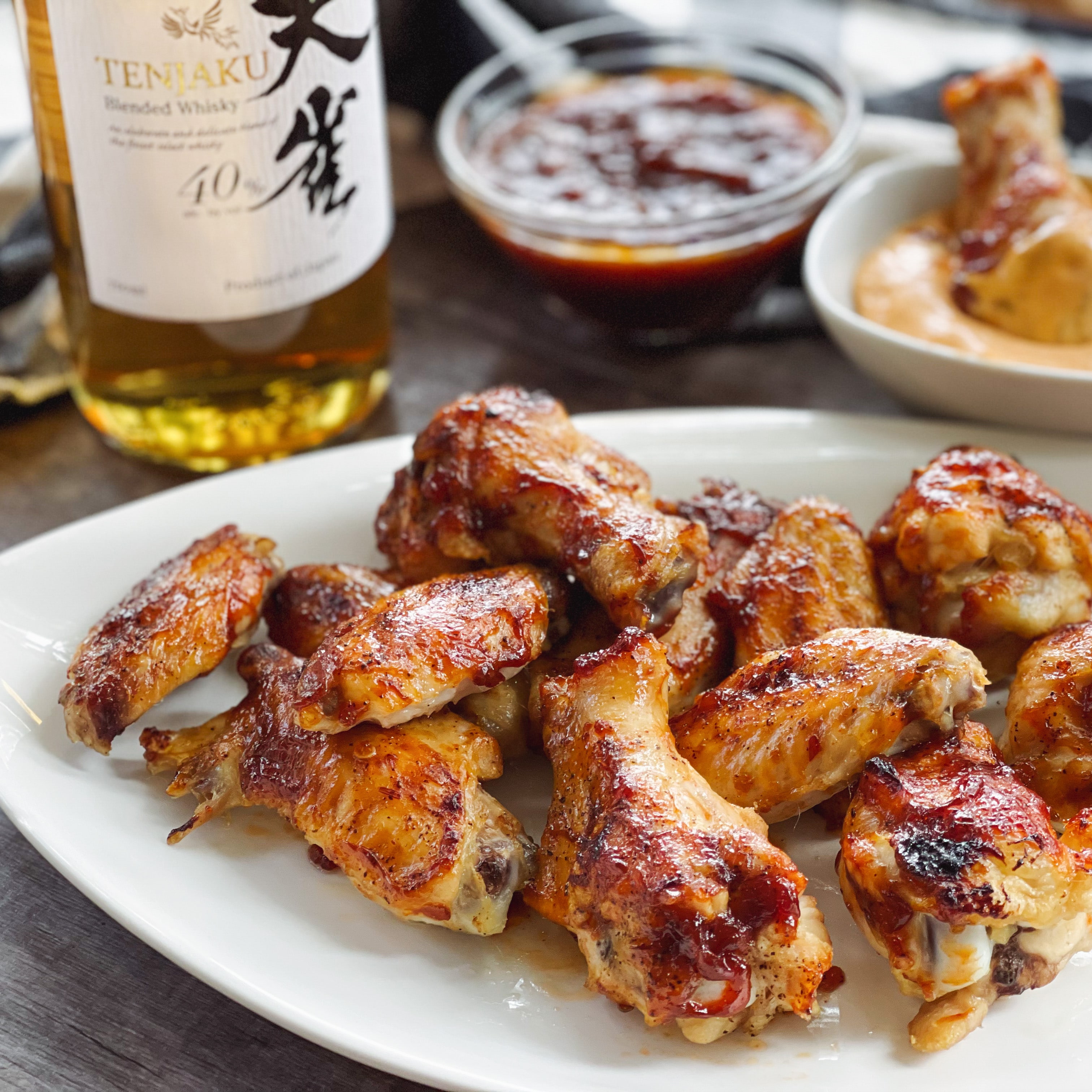 A bottle of Japanese whiskey placed behind a white plate of saucy chicken wings, celery and carrot sticks, with a bowl of aioli dip and extra sauce behind.