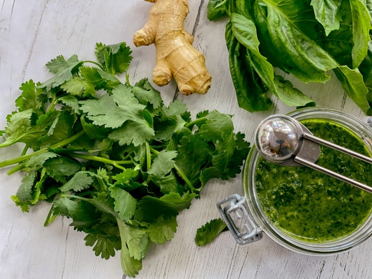 Cilantro, basil, and ginger root on a white board with a bowl of green sauce on the side.