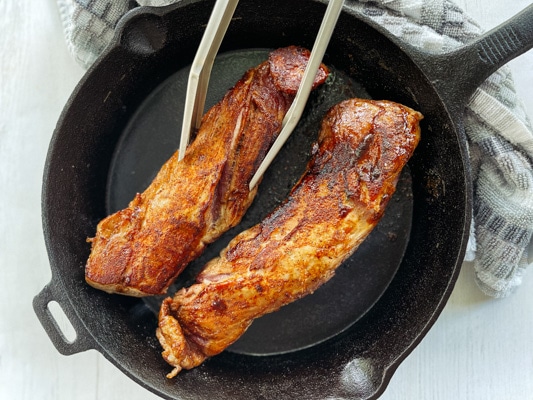 Seared and caramelized pork tenderloin filets in a cast iron pan on top of a white board.