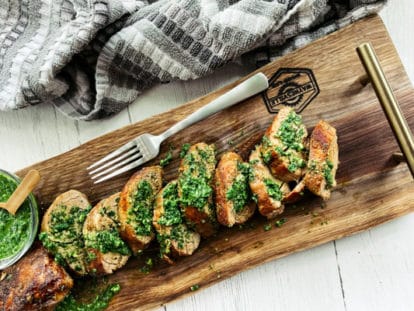 Thick slices of pork tenderloin on a rustic wooden board topped with a vibrant green cilantro, mint, basil sauce and a small bowl of sauce and a fork on the side.