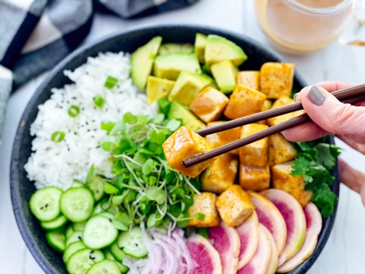A woman holding chopsticks with a tofu cube, and a miso tofu poke bowl underneath.
