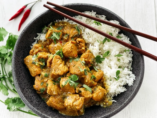 A black bowl of Thai curry chicken with white rice and chopsticks with fresh cilantro and red chili peppers on the side, on top of a white board.