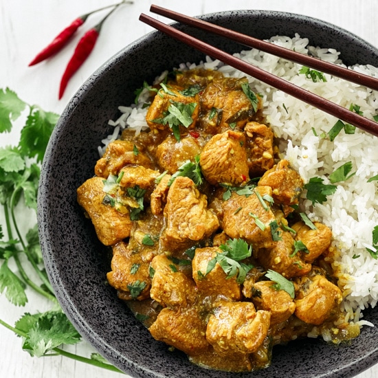 A black bowl of Thai curry chicken with white rice and chopsticks with fresh cilantro and red chili peppers on the side, on top of a white board.