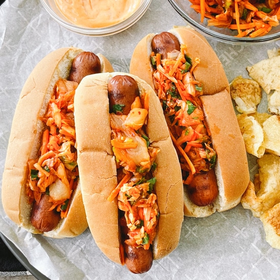 Three hot dogs in buns topped with kimchi slaw with potato chips, extra slaw and aioli on the side.