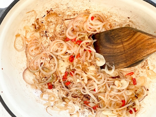 Shallots and Thai chili peppers being sauteed in a large Dutch oven with a wooden spatula.