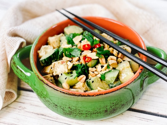 A green bowl filled with a vibrant spicy Asian tofu salad with cucumbers, cilantro and peanuts, and black chopsticks on top.